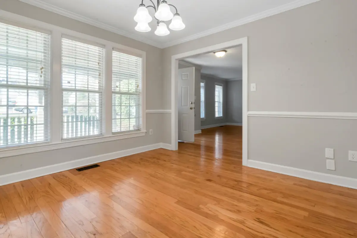 Bright, updated room with a freshly renovated wooden floor, light gray walls, large windows with blinds, hanging light fixture, and an open doorway leading to another room.