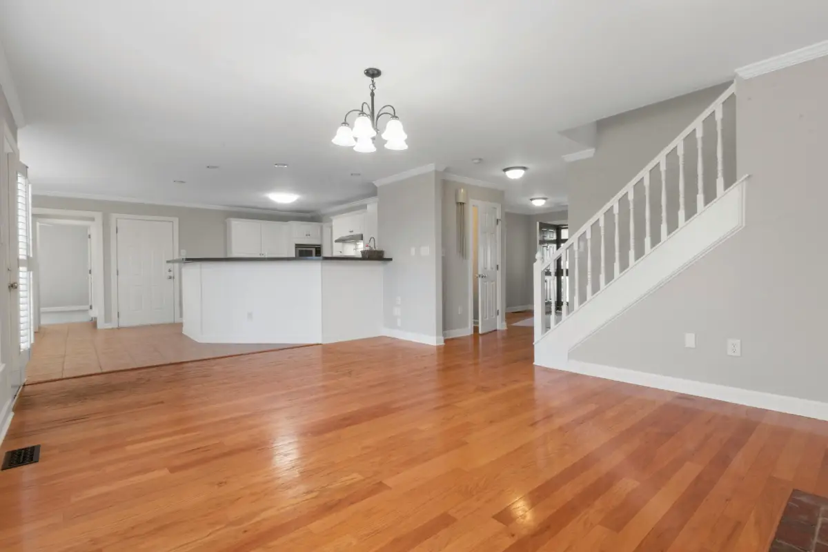 A brightly lit interior of an unfurnished home with newly completed floor renovation showcases wooden floors, a white kitchen island, white walls, and a staircase leading to the upper floor. A ceiling light fixture is centered in the living area.