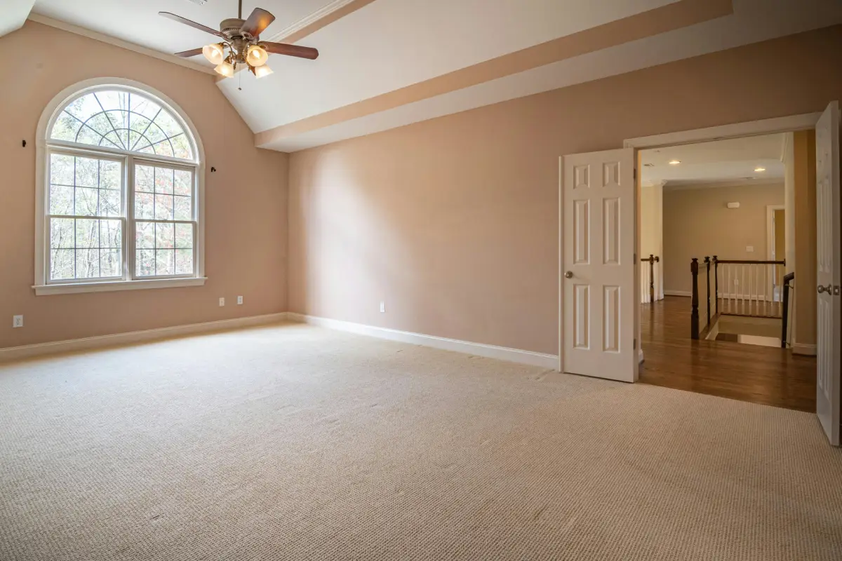 An empty, carpeted room with beige walls, a high ceiling, and a ceiling fan stands ready for floor renovation. The space features an arched window and a door that opens to a hallway with wooden flooring.